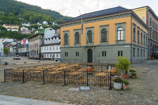 View of a historic building with a yellow facade stands proudly on a cobblestone square in Bergen, Vestland, Norway, with rows of wooden tables.