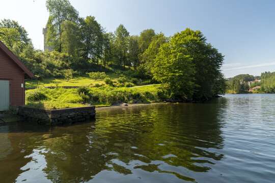View of the tranquil lake mirroring the lush green hillside crowned with a stone structure, the red boathouse adding a touch of rustic charm, Bergen, Vestland, Norway.