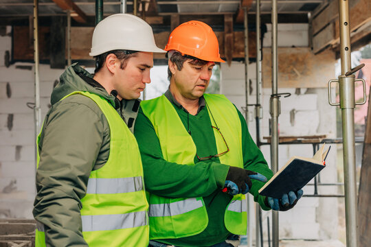 Builder in hard hat checking construction plan with notebook outdoors