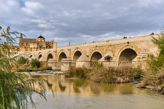 View of the ancient Roman bridge stretches across the Guadalquivir River, leading to the Mezquita-Cathedral under a cloudy sky, Cordoba, Andalusia, Spain.