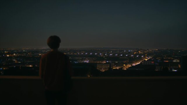 Gazing person leaning on parapet on roof at night, shoulder bag and jacket, city lights brightening