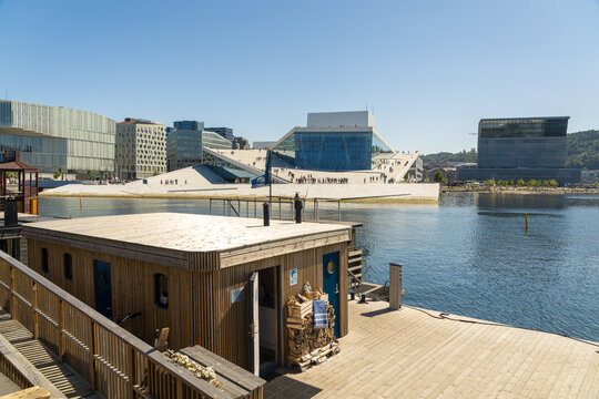 View of wooden structure with a flat roof and blue trim, alongside the modern architecture of the Oslo Opera House, reflecting in the water, Oslo, Oslo, Norway.