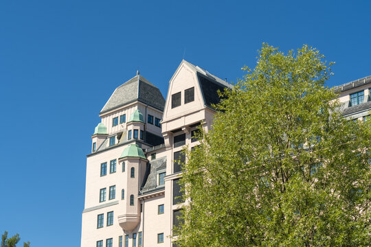 View of a building with light pink walls and green turrets contrasts against the bright blue sky, partially obscured by vibrant green leaves, Oslo, Norway.