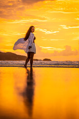 Fototapeta premium Woman Walking Along Reflective Wet Sand, Soft Backlight From Low Sun, Gentle Waves, Tote Bag Swing, Contemplative Mood, Warm Golden Palette, Long Shadow Cast, Travel Lifestyle Scene