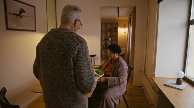 Tracking shot of caring senior Caucasian man bringing plates and serving meal for his African American wife working with documents on laptop at table and having dinner together in modern apartment