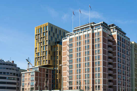View of modern buildings rise against a clear blue sky, flags fluttering atop one, a blend of golden and brick hues, Oslo, Oslo, Norway.