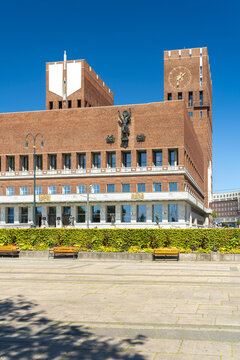 View of the imposing brick facade of Oslo City Hall, juxtaposed against a vibrant blue sky, rising above manicured greenery, Oslo, Oslo, Norway.