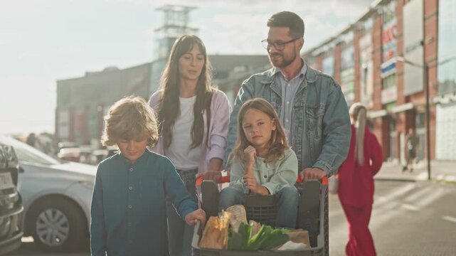 Parents with two children walking after shopping. Girl sitting in grocery cart with food bags, boy walking beside, mother and father looking at each other. Family outdoors near shopping center.