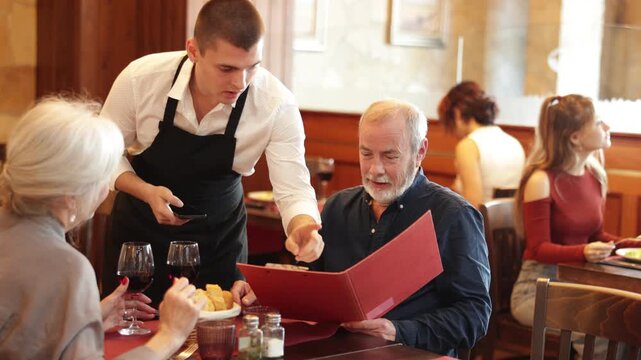 Polite waiter taking order from elderly couple during relaxed dinner in comfortable restaurant setting. High quality 4k footage