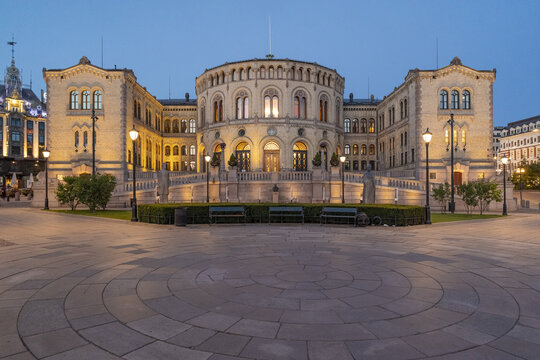 View of the Storting building illuminated against the twilight sky, contrasting with the patterned stone plaza, creating a serene atmosphere, Oslo, Oslo, Norway.