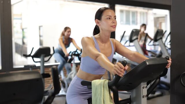 Positive young woman doing spin bike exercises in well-equipped gym during training session