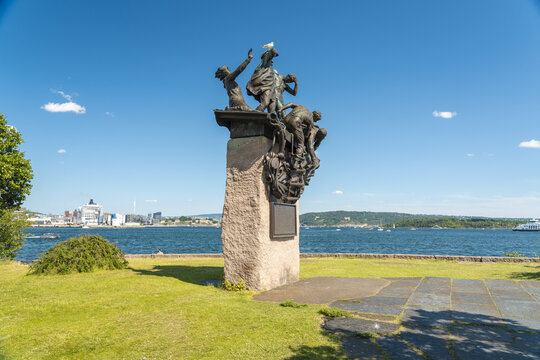 View of a bronze sculpture on a stone pedestal stands in stark contrast against the vibrant blue sea and sky, framed by green grass, Oslo, Oslo, Norway.