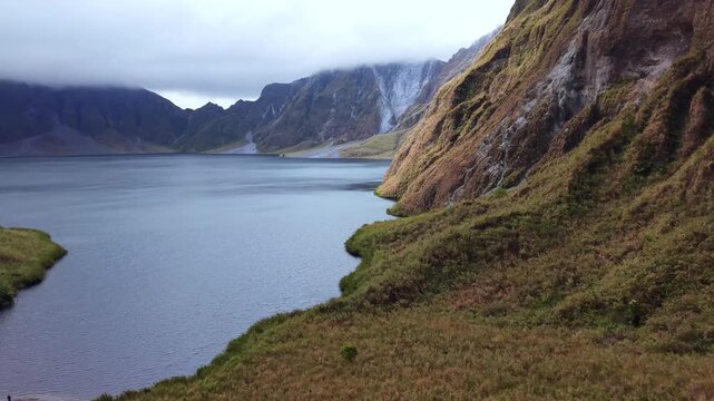 Pinatubo, philippines. Calm lake reflects distant mountain peaks. Lush grass slopes lead down to the water's edge. Hiker walks the path with quiet focus. Their calm demeanor suggests deep appreciation