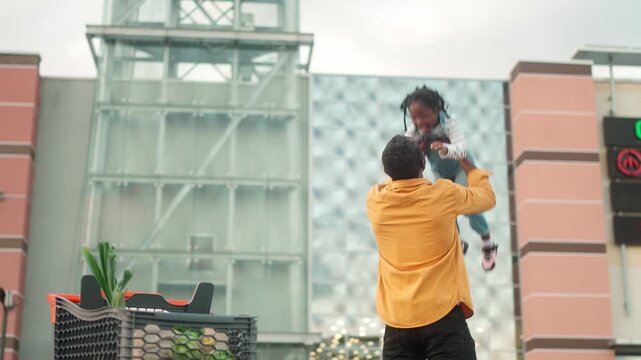 African American young father holding daughter standing near supermarket and tossing up. Male parent lifting child in arms outdoors with shopping cart full of groceries in parking lot.