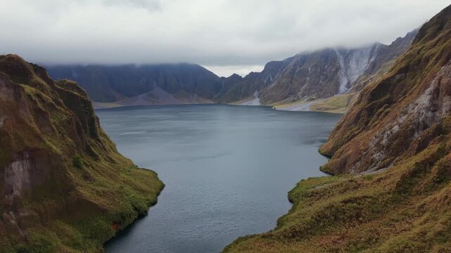 Pinatubo. Deep blue lake reflects cloudy sky. Rugged mountains frame calm waters. Lush grasses cover slopes with patches of wildflowers. Fog drifts over distant peaks