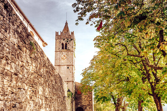 View of an ancient stone wall leading to a clock tower framed by autumn foliage, showcasing a rich blend of history and natural beauty, Hum, Croatia.