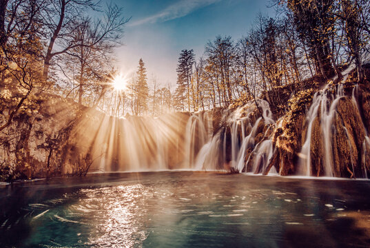 View of sunbeams filtering through bare trees illuminating cascading waterfalls flowing into a serene pool of water, Plitvicka Jezera, Croatia.