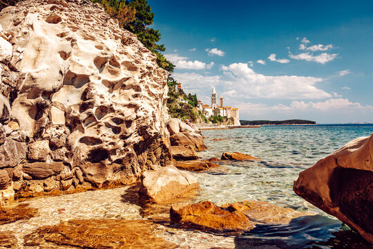 View of rugged, sun-kissed rocks meet the turquoise sea, leading the eye towards the distant white buildings under a bright sky, Rab, Primorje-Gorski Kotar County, Croatia.