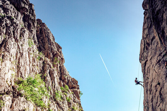View of a climber rappelling down a steep rock face, with a jet stream slicing through the clear blue sky, framed by immense cliffs, Starigrad, Croatia.