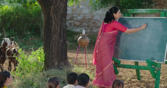 Indian mature adult teacher do study work teach to villager young girl boy rural area. Happy child group sitting on mat look board learn English enjoy desi life open class day time field ground place