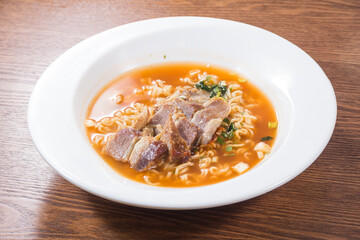 A white bowl of ramen noodles in spicy broth topped with sliced pork and vegetables served on a wooden table.