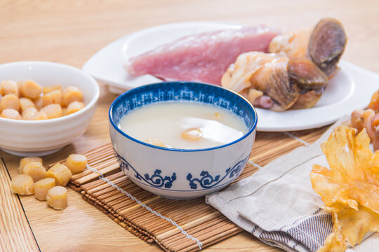 A ceramic bowl of white soup placed on a wooden table surrounded by dried scallops, pork, and mushrooms.