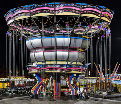 View of a vibrant carousel, its lights ablaze against the night sky, casting shadows on the wet ground in La Spezia, Liguria, Italy.