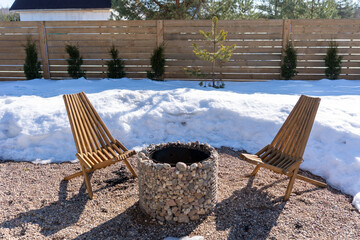 Modern outdoor fire pit area in winter featuring two wooden slatted chairs and a stone gabion...