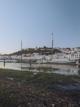 View of white buildings with terracotta roofs cascading down the hillside towards the tranquil waters reflecting the serene sky, Alcacer do Sal, Setubal, Portugal.