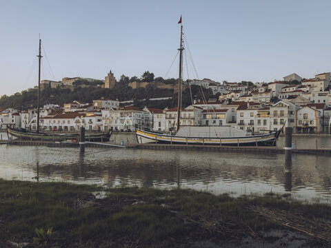 View of traditional boats rest on calm waters reflecting the golden light of dusk against the backdrop of white buildings, Alcacer do Sal, Setubal, Portugal.