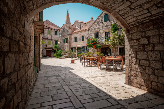 View of sun-drenched stone buildings under a clear blue sky, framed by a rustic stone archway, casting shadows on the cobblestone ground, Stari Grad, Croatia.