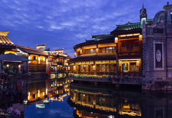 Obraz premium Night view of traditional stilt houses with glowing lanterns reflecting on the river in Moon Fields (Yueliangtian) scenic spot in Lizhuang Ancient Town, Yibin, Sichuan, China