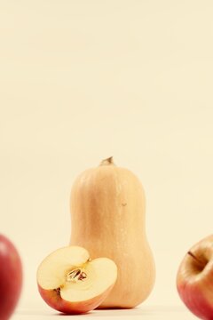 Vertical macro of butternut squash with halved red apple on pastel yellow background. Blurry whole red apples in foreground. Fresh organic fruits and vegetables with copy space on top for text.