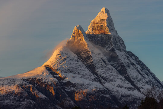 View of a majestic, snow-dusted mountain peak ablaze with the warm hues of the setting sun, contrasting against the cool, serene blue sky, Skibotn, Norway.