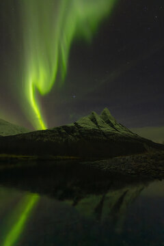 View of emerald auroras dance above the dark, jagged peaks mirrored in the tranquil waters below, casting an ethereal glow over the landscape, Skibotn, Troms, Norway.
