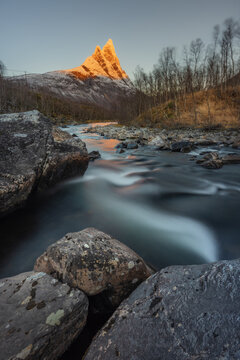 View of a glacial river rushing past moss-covered boulders towards a towering, sunlit peak amidst bare trees, creating a serene yet dramatic landscape, Skibotn, Norway.