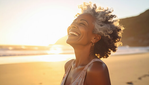 Mujer senior de mas de 50 a&ntilde;os, afroamericana con pelo rizado corto y canoso, sonriendo con los ojos cerrados con fondo borroso de una puesta de sol a la orilla del mar en una playa vacia