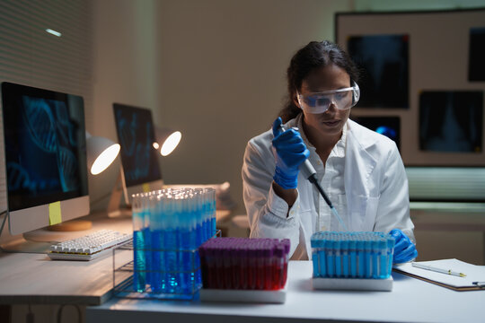 Young female scientist performing an experiment with a micropipette and test tubes, analyzing DNA sequences on a computer screen in a dark laboratory for medical and genetic research