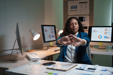 Businesswoman stretching at her desk in a dim office after overtime, rubbing tired shoulders amid...