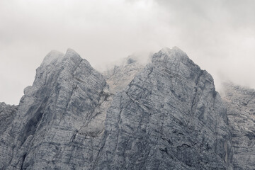 A somber, high-contrast view of jagged limestone mountain faces in the Julian Alps partially obscured by thick, low-hanging clouds on a misty day. Black and white textured stock photo