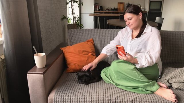 Woman sitting on sofa with small griffon dog, using smartphone enjoying quiet moment at home. Concept of slow living lifestyle, relaxation, cozy atmosphere, and peaceful everyday routine with pets.