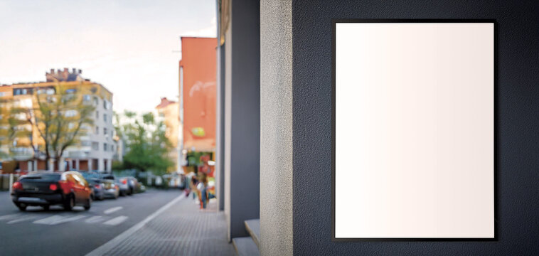 Blank vertical poster mockup in a black frame on a textured city wall with a blurred traffic background.