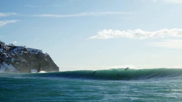 Powerful ocean swell rolling toward surfers near rugged coastal cliffs, low in water slow motion perspective