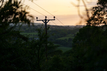 Naklejka premium Rural landscape in Wales with power lines against a warm sunset. Golden light highlights rolling hills and countryside contours.