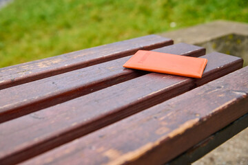 Fototapeta premium Orange passport holder forgotten on weathered wooden park bench outdoors