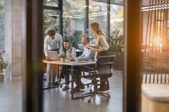 A view through a glass partition shows a modern, busy office interior. Business people are working at desk.