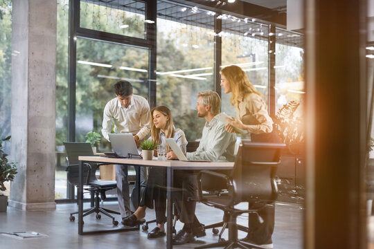 Group of business people standing together and discussing their work and projects, having a team meeting in an office, view from glass wall.