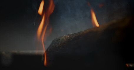 Fototapeta premium Low key close up of orange flames and smoke behind a dark log, creating a moody fire background for heat, danger, barbecue, and atmospheric concepts.