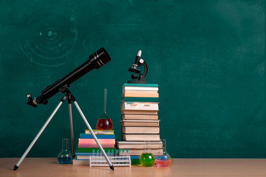 Natural sciences classroom scene with telescope, textbooks, microscope and chemistry glassware against green chalkboard. Blank space for text.