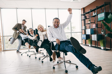 Obraz premium Group of colleagues engaging in joyful office activity, sliding on chairs in open workspace, exuding teamwork and camaraderie